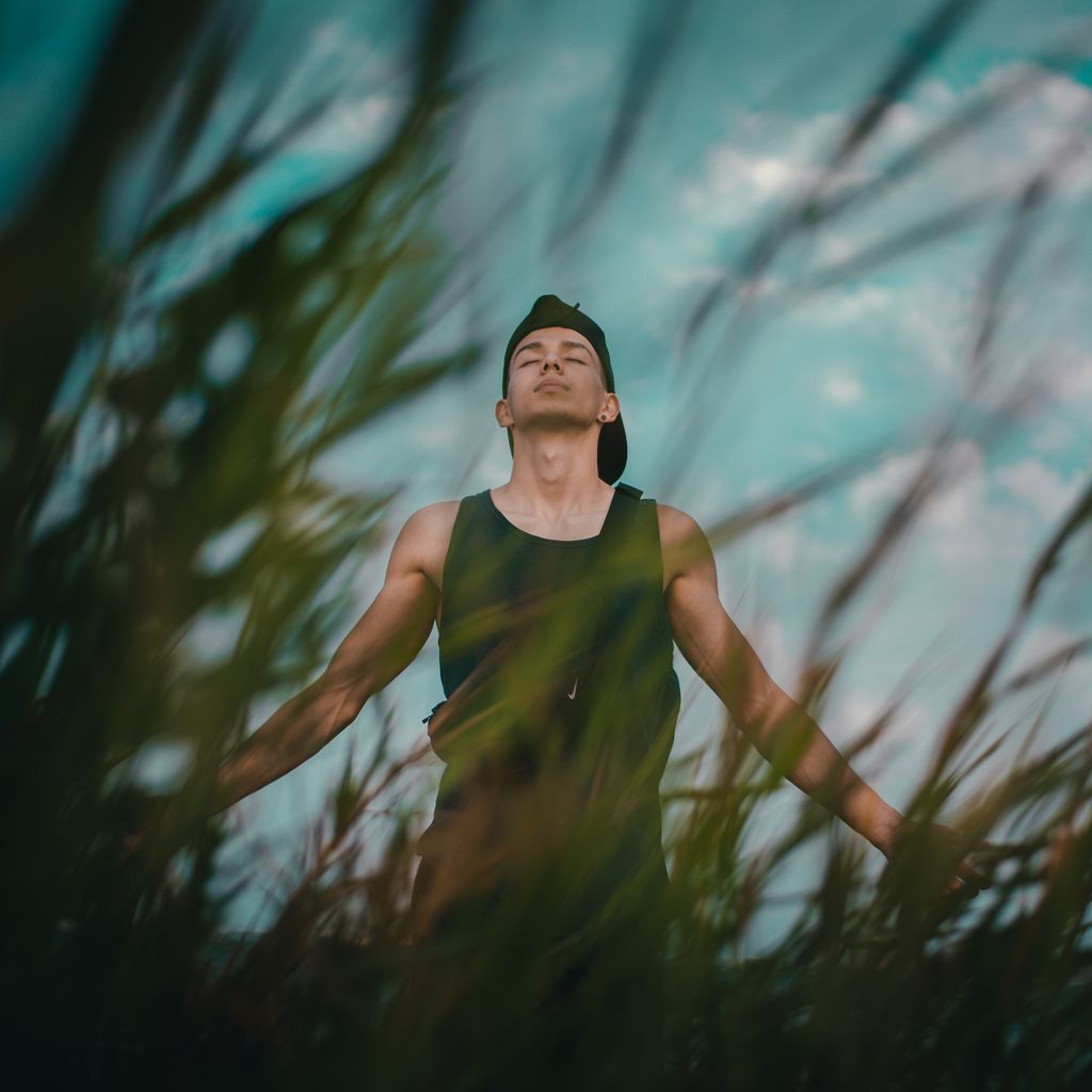 A young man relaxes and meditates among tall grass outdoors, eyes closed, embracing nature.
